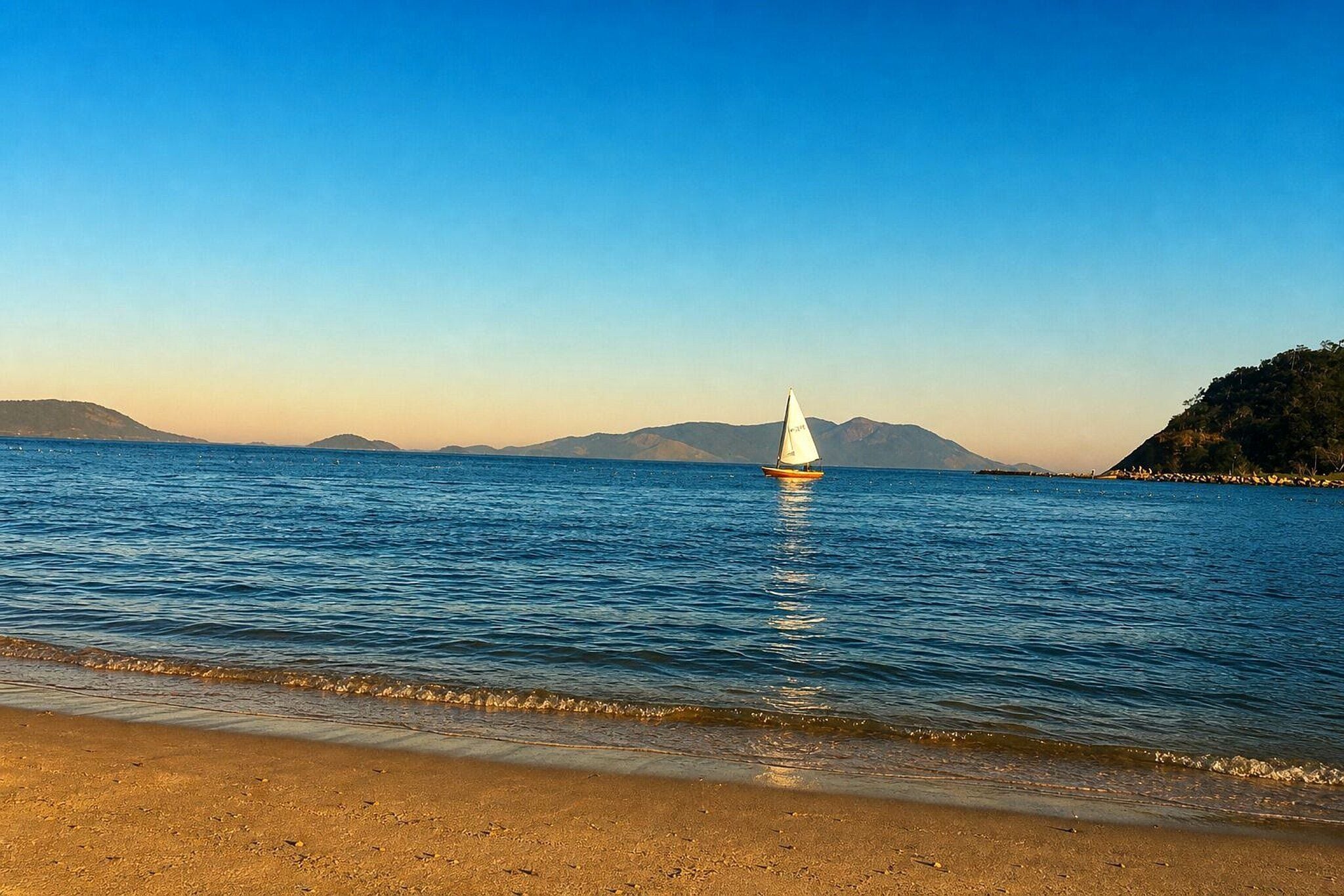 A beach with a sailboat on calm waters, a clear sky, and distant mountains.