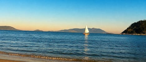 Una playa con un velero en aguas tranquilas, cielo despejado y montañas a lo lejos.