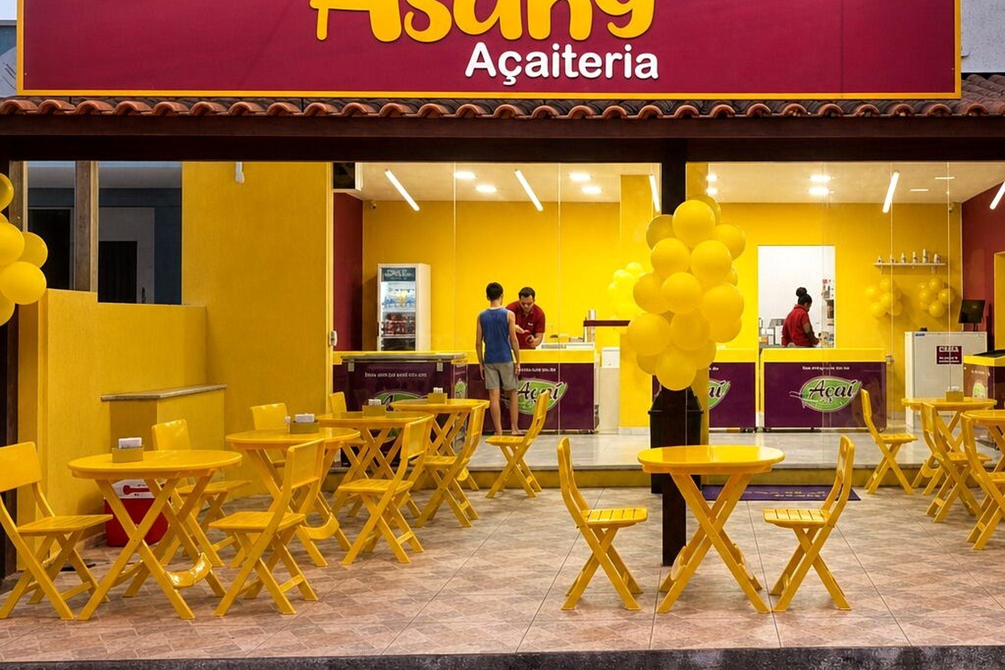 A café with yellow chairs and tables, a counter with a person behind it, and a sign above reading 'Açaieria.'