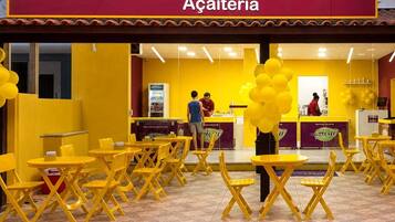 A café with yellow chairs and tables, a counter with a person behind it, and a sign above reading 'Açaieria.'