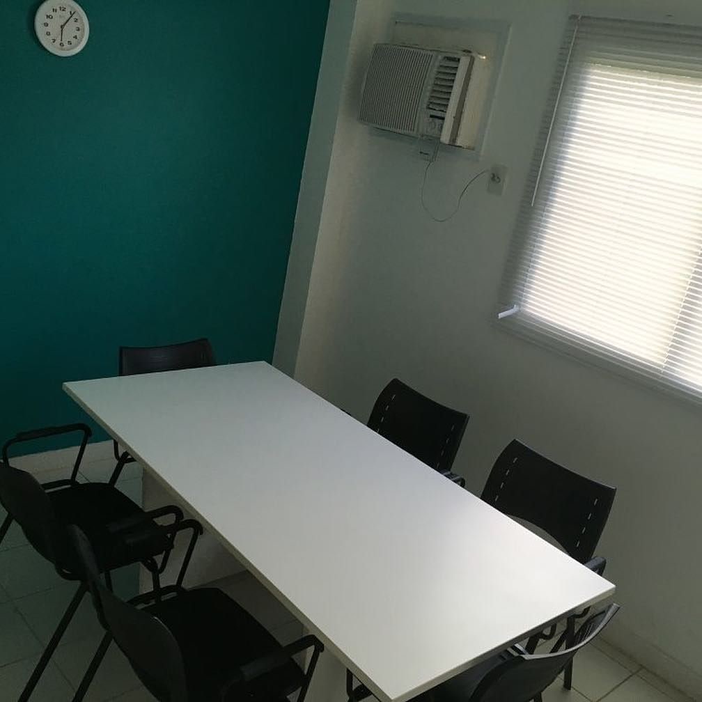 Uma sala de conferências com uma mesa branca, cadeiras pretas, um relógio na parede, um aparelho de ar condicionado e uma janela com persianas.