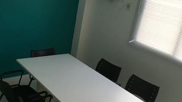 Uma sala de conferências com uma mesa branca, cadeiras pretas, um relógio na parede, um aparelho de ar condicionado e uma janela com persianas.