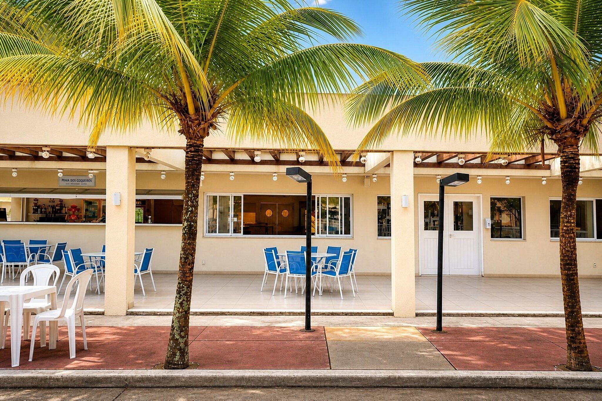 An outdoor seating area with tables and chairs, palm trees, and a building with large windows.
