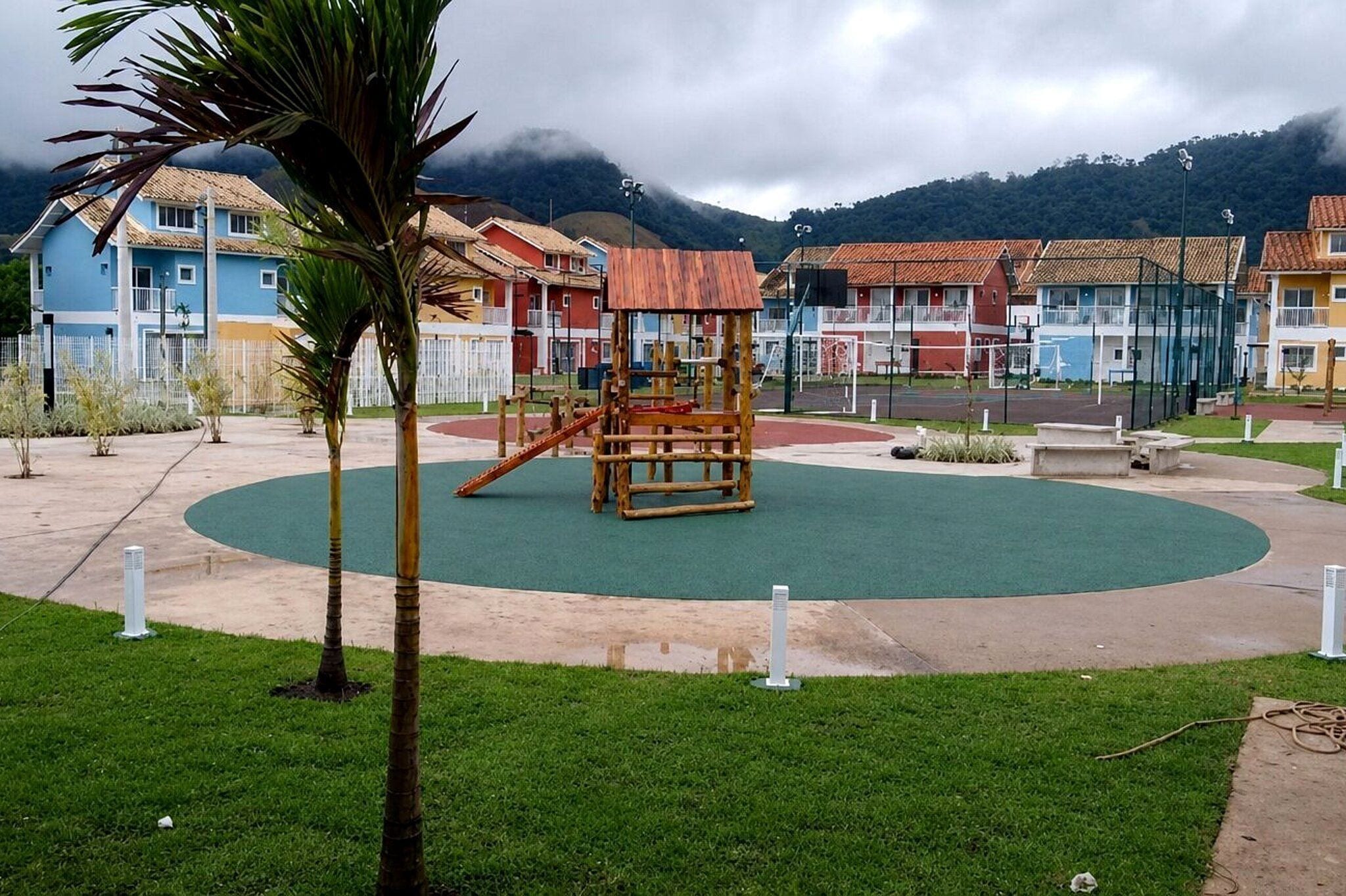 A playground with a wooden play structure, surrounded by a green field and a paved area with buildings in the background.
