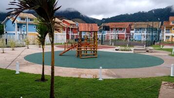 A playground with a wooden play structure, surrounded by a green field and a paved area with buildings in the background.