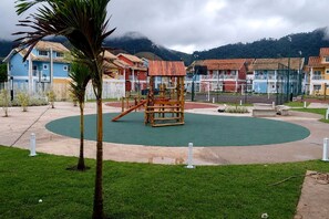 A playground with a wooden play structure, surrounded by a green field and a paved area with buildings in the background. - Reserva do Sahy, Condado (Mangaratiba)