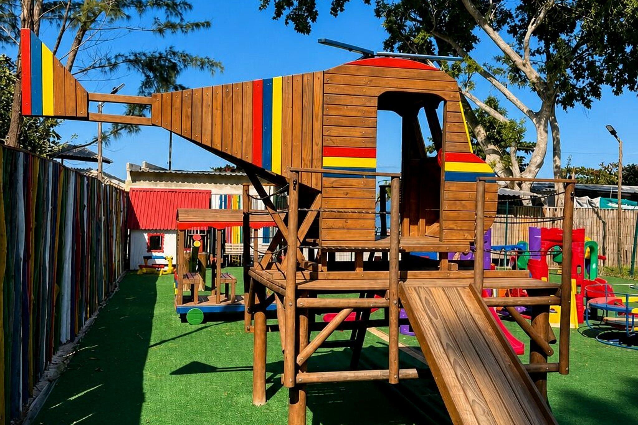 Une structure de jeu en bois, dotée d’un toboggan, entourée d’une zone délimitée par une clôture et agrémentée de décorations colorées.