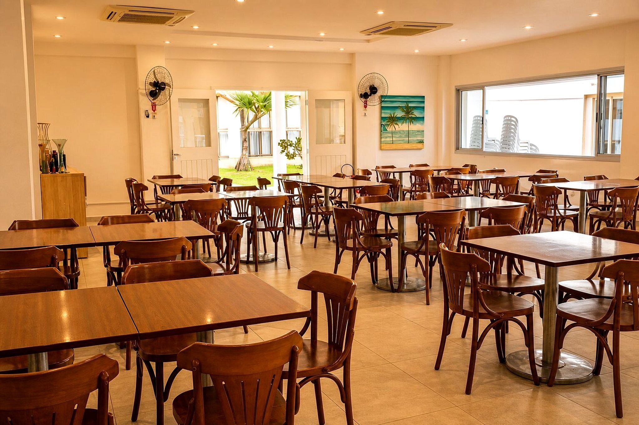 A spacious dining area with multiple wooden tables and chairs, large windows offering a view of the city, and ceiling fans.
