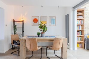 A dining area with a wooden table, two chairs, a potted plant, and framed artwork on the wall.