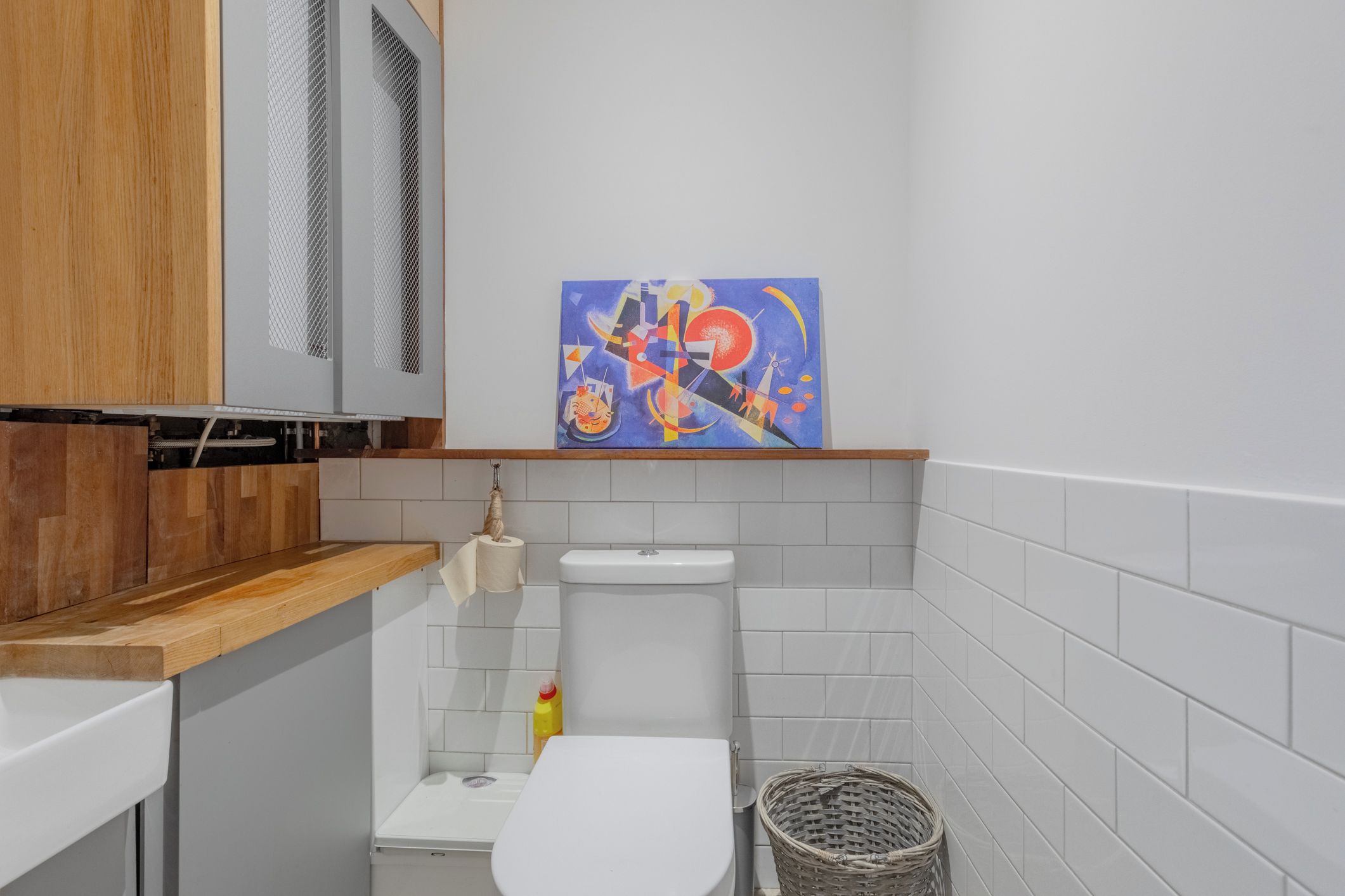 Een compacte badkamer met een wit toilet, een klein wastafeltje en een houten aanrechtblad. Een schilderij hangt aan de muur boven het toilet.