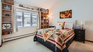 A bedroom with a large bed, a window with blinds, a bookshelf, and a bedside table. - Stone Creek Meadows 205 2 Bedroom Condo (Avon)
