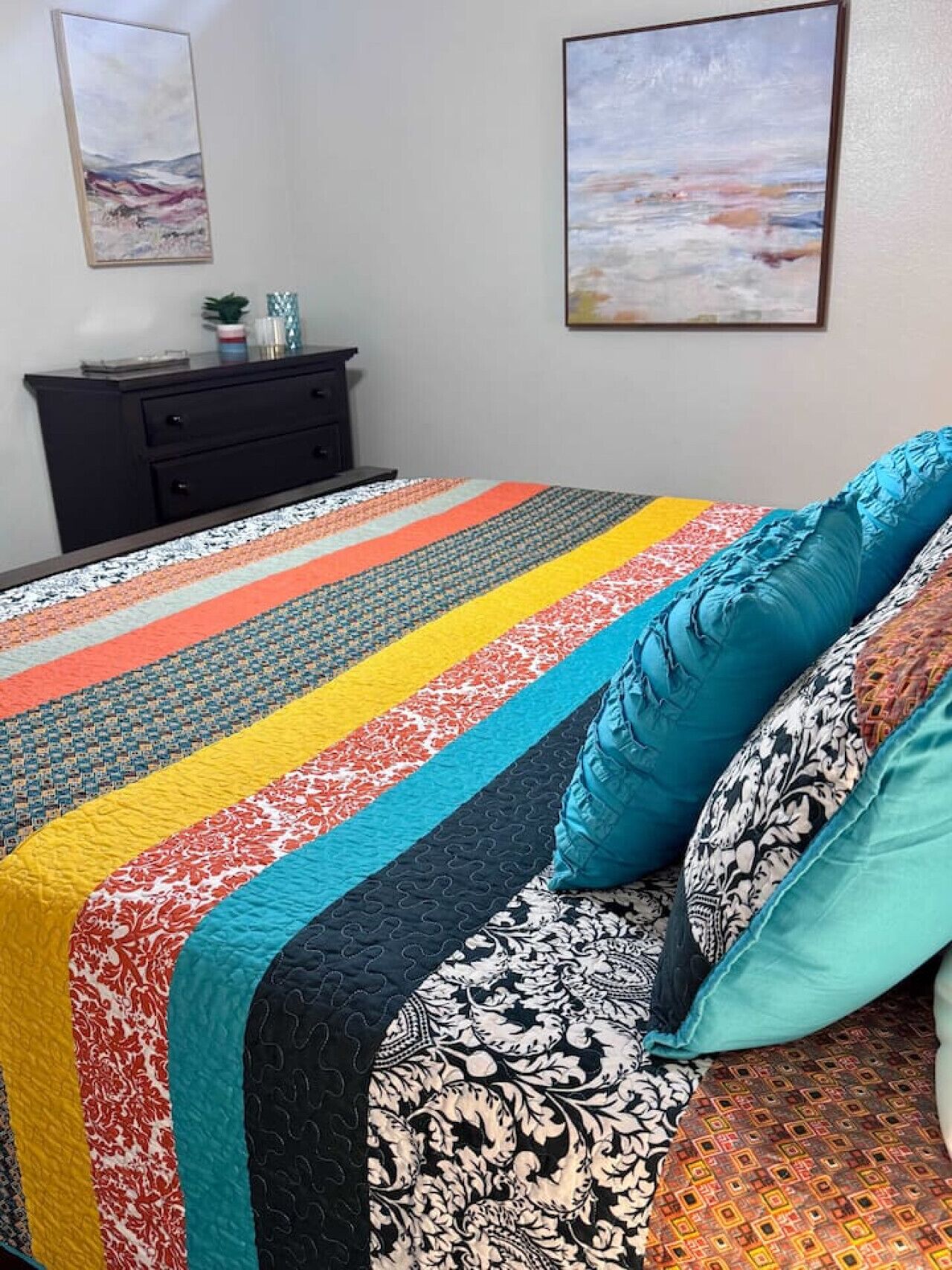 A bed with a colorful quilt and patterned pillows, a black dresser, and two framed artworks on the wall.