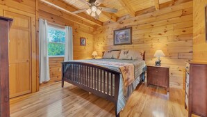 A wooden cabin bedroom with a bed, two bedside tables, a ceiling fan, and a window with curtains. - Chillin N Grillin 2 Bedroom Cabin (Sevierville)
