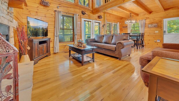 A spacious wooden cabin with a loft bedroom, a fireplace, a dining area, and a large window overlooking the forest. - Chillin N Grillin 2 Bedroom Cabin (Sevierville)