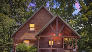 A wooden cabin with a porch and two windows, surrounded by trees. - Chillin N Grillin 2 Bedroom Cabin (Sevierville)