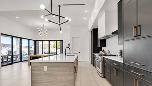 A modern kitchen with dark cabinetry, a central island, and a dining area with large windows. - Luxury Lounge - 5BR, Pool&Spa, Sauna, Firepit (Scottsdale)