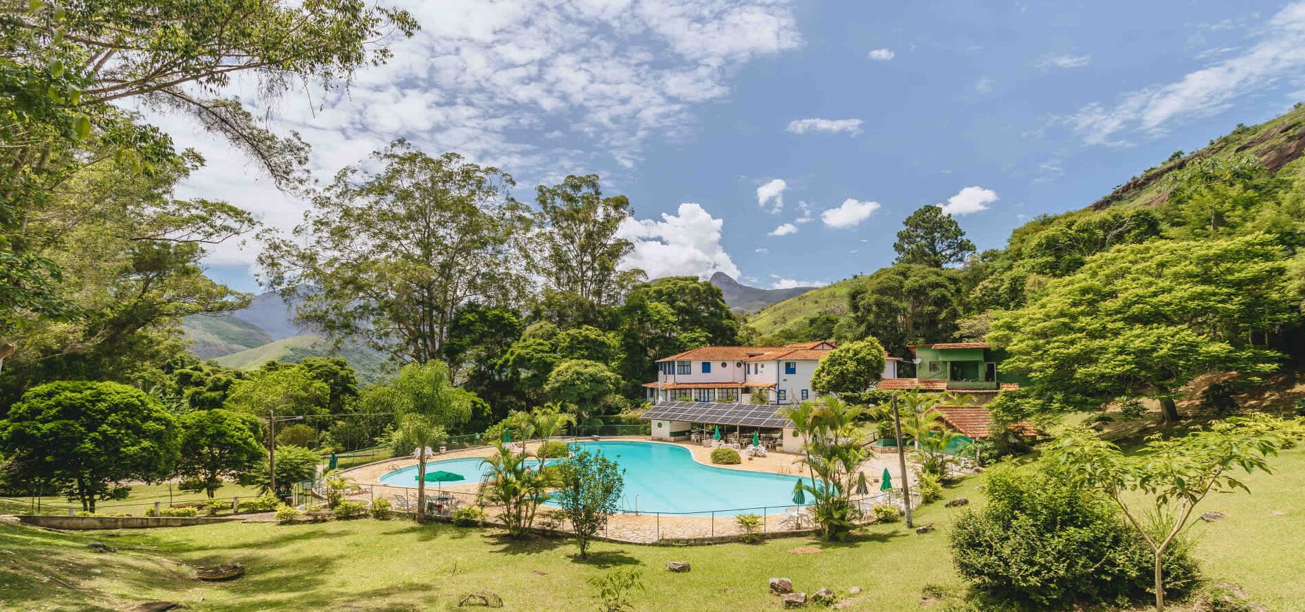 Piscina rodeada por vegetação exuberante e uma casa com telhado vermelho.