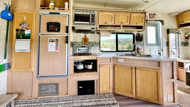 A compact RV kitchen with wooden cabinets, a refrigerator, microwave, toaster, sink, and a window offering a view of the outdoors.