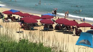 Une plage avec des parasols rouges, une jetée en arrière-plan, et des gens qui nagent et prennent des bains de soleil.