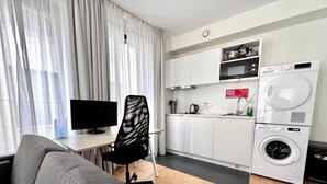 Un espacio habitable pequeño con sofá, una silla de oficina tipo malla negra, televisor de pantalla plana, lavadora y kitchenette con armarios y pileta.