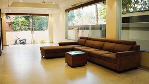 A spacious room with a large sectional sofa, a glass coffee table, and a view of the outdoors through large windows. - ARK Hotels (Ranchi)