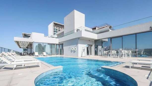 Ein Pool auf dem Dach mit Liegestühlen, ein modernes Gebäude mit großen Fenstern und ein klarer Himmel.