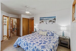 A bedroom with a bed, a ceiling fan, a nightstand, a lamp, and a painting of a beach scene.