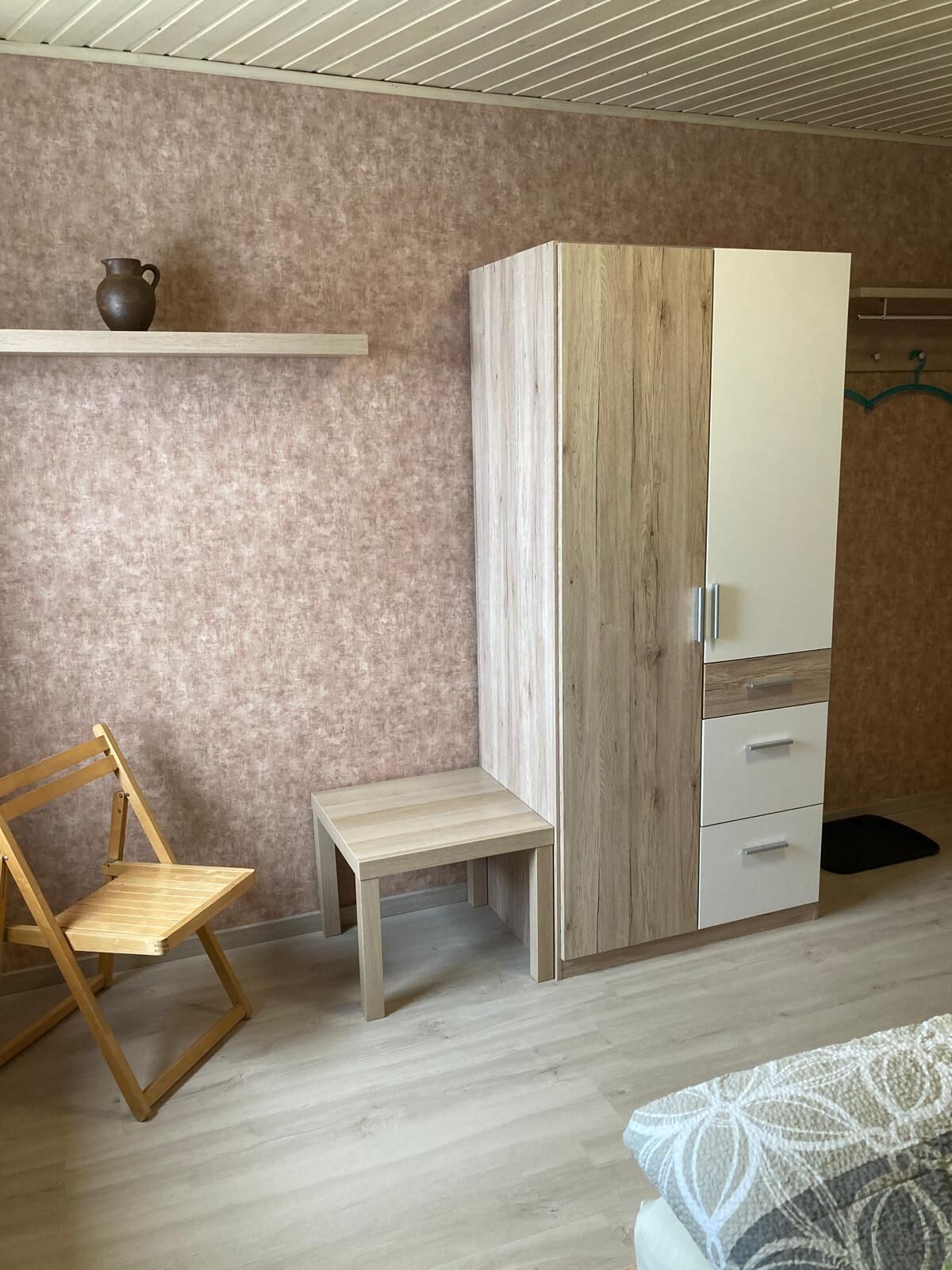 Een slaapkamer met een houten garderobe, een klein houten tafeltje, een stoel en een plank met een pot.