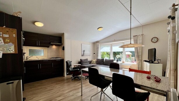 A modern kitchen with dark cabinets, a wooden countertop, and a built-in oven. There is a dining area with a table and chairs, a sofa, and a window with a view of greenery outside.