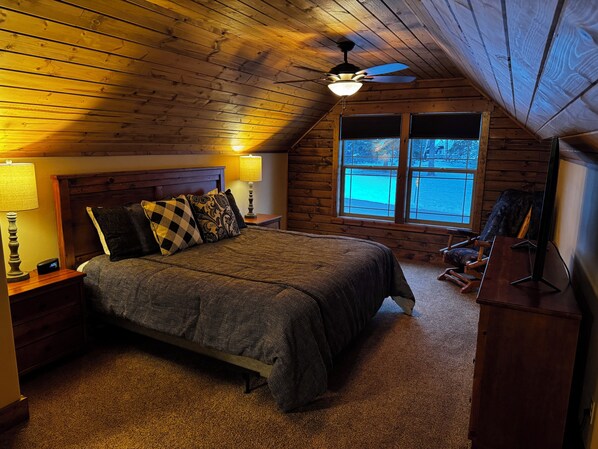 Une chambre confortable dans un chalet en rondins, avec un lit, une table de chevet, une lampe, un ventilateur de plafond et une fenêtre donnant sur un paysage enneigé.