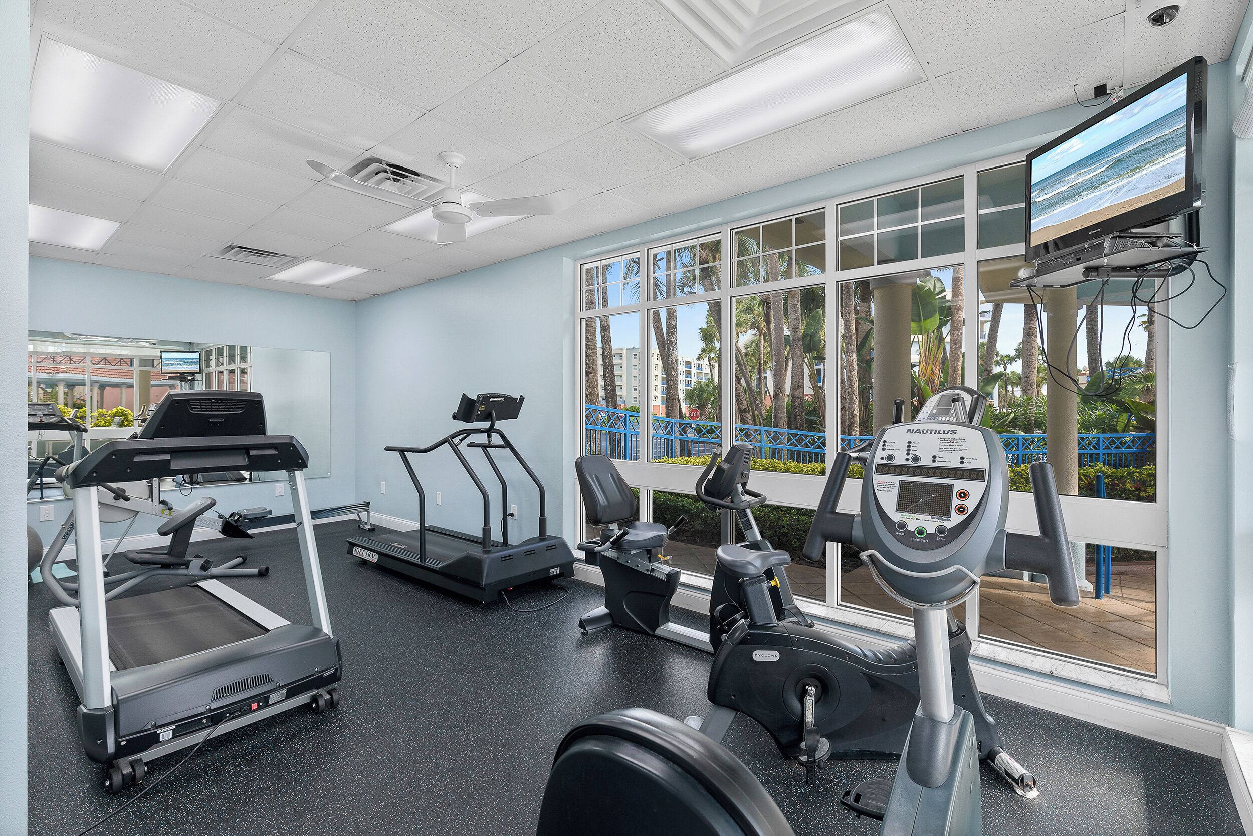 A gym with treadmills, elliptical machines, and a stationary bike, large windows with a view of palm trees and buildings.