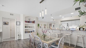 Una cocina moderna con armarios blancos, una isla central con taburetes y un área de comedor con mesa de vidrio y sillas de mimbre.