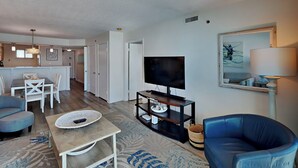 A modern living room with a TV, seating, and a kitchen in the background. - Sea Oats #603 | Gulf Front Condo w/ Pool + Gym and Near Dining + Activities (Fort Walton Beach)