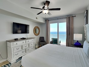 A hotel room with a bed, a TV, a ceiling fan, a dresser, a chair, a lamp, and a view of the ocean.