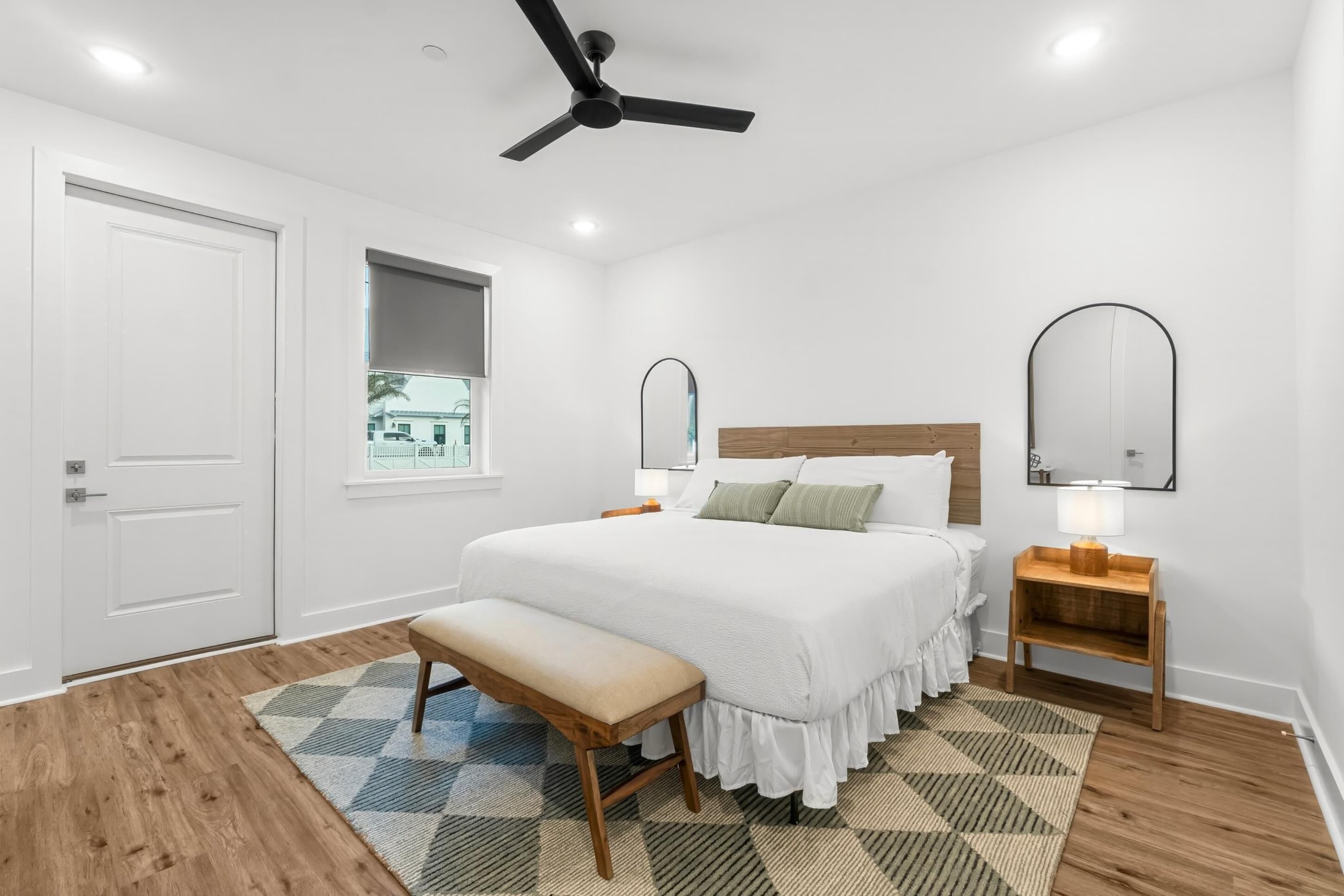 A bedroom with a bed, a bench, a ceiling fan, a window, and a nightstand with a lamp.