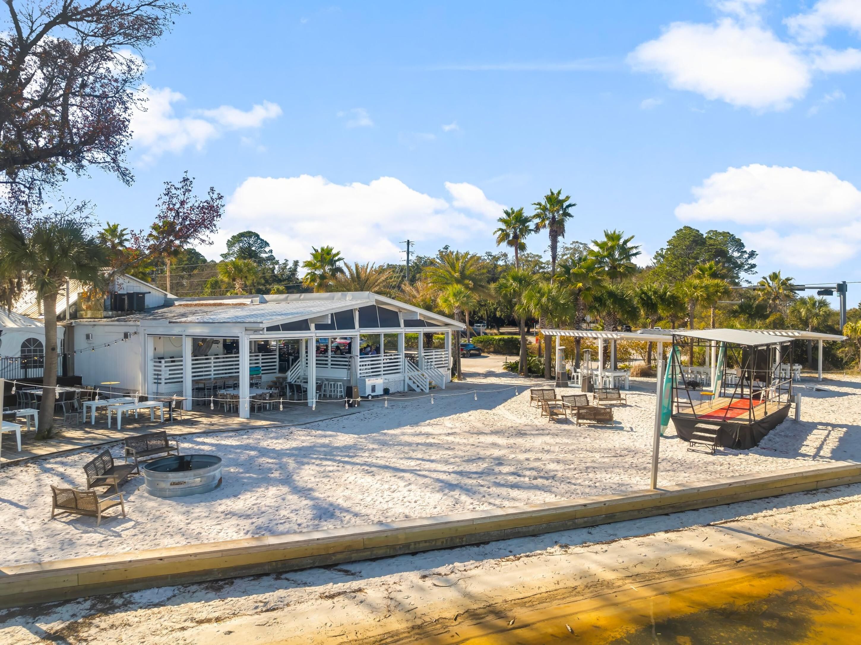 A beachside restaurant with outdoor seating, a covered area, and a boat on the sand.