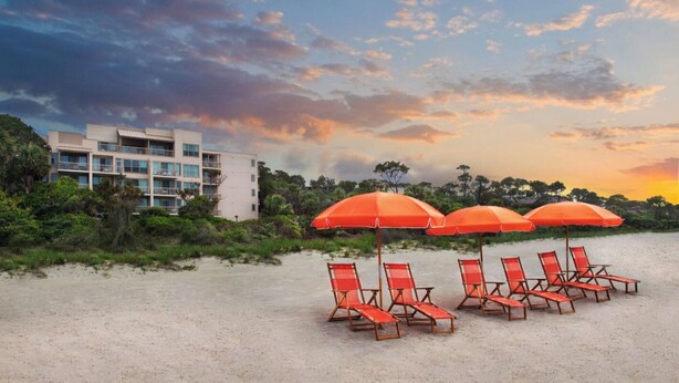 A beach with several orange umbrellas and lounge chairs, a modern building with balconies, and a sunset sky.