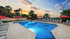 A swimming pool with lounge chairs and umbrellas, surrounded by trees and a sunset view.