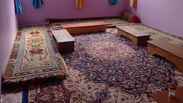 A room with a purple wall, a patterned carpet, and a bench along the window. - Lammaney Homestay (Lahul)