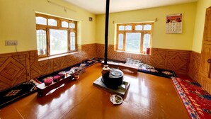 A room with wooden flooring, two windows with wooden frames, a central heating stove, and a patterned rug. - Lammaney Homestay (Lahul)