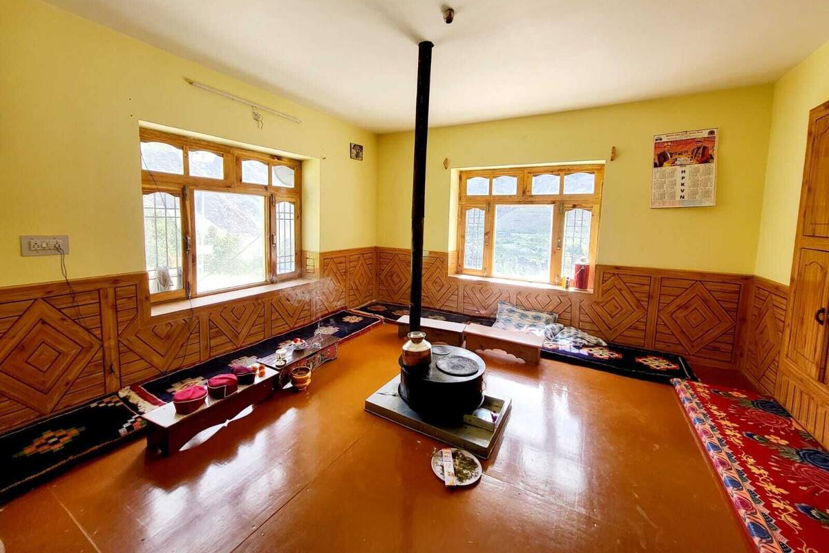 Una habitación con suelo de madera, dos ventanas con marcos de madera, un estufa de calefacción central y una alfombra estampada.