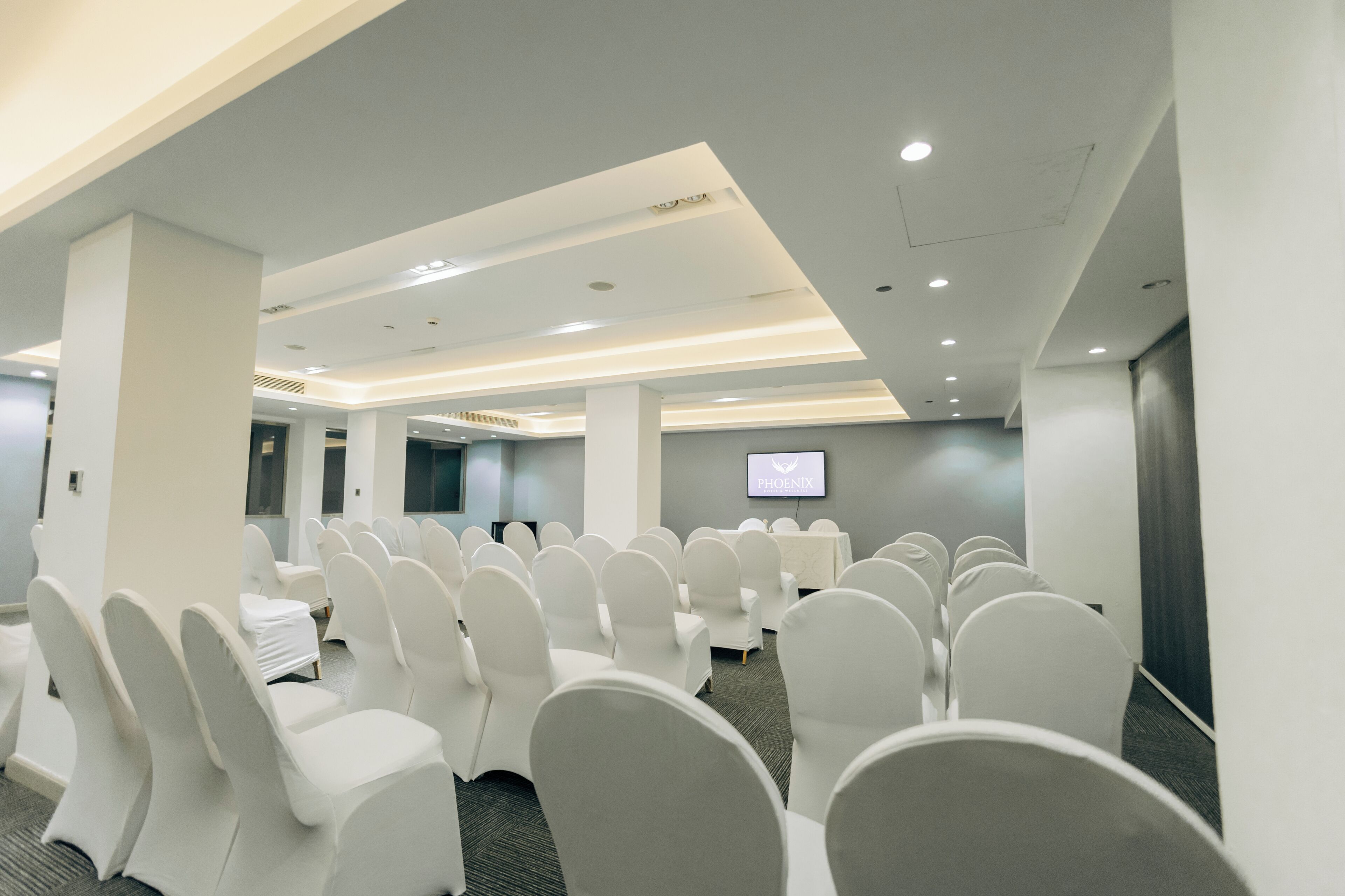 Un salón de conferencias con filas de sillas blancas, un televisor de pantalla plana en la pared e iluminación empotrada en el techo.
