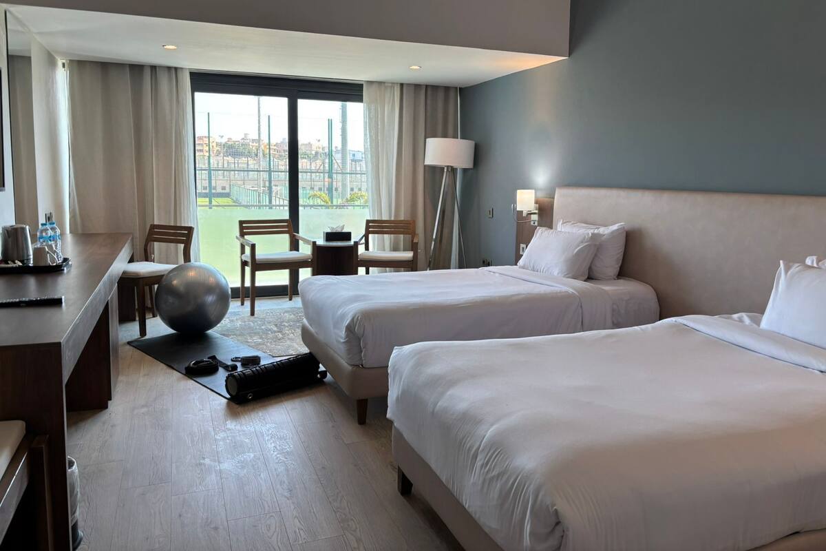 A hotel room with two beds, a desk, a chair, a lamp, and a view of the city.