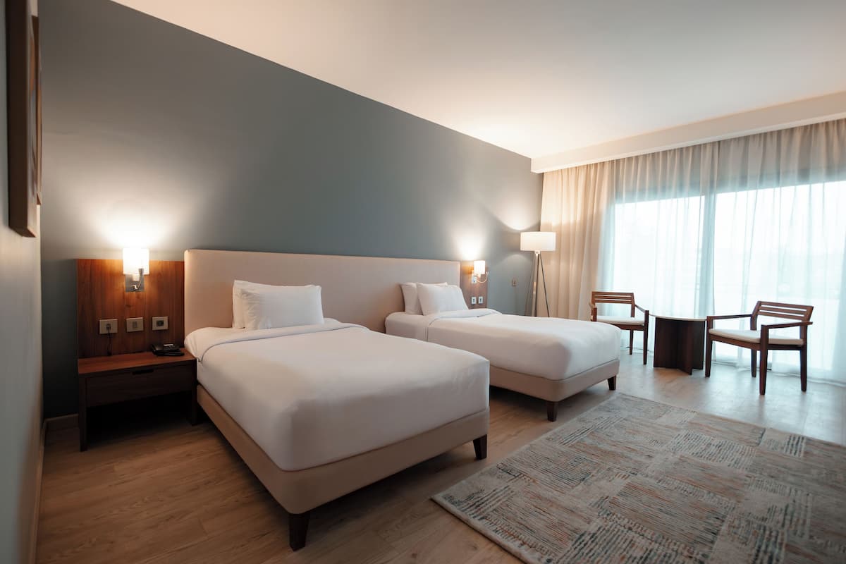 A hotel room with two beds, a desk, chairs, and a large window.