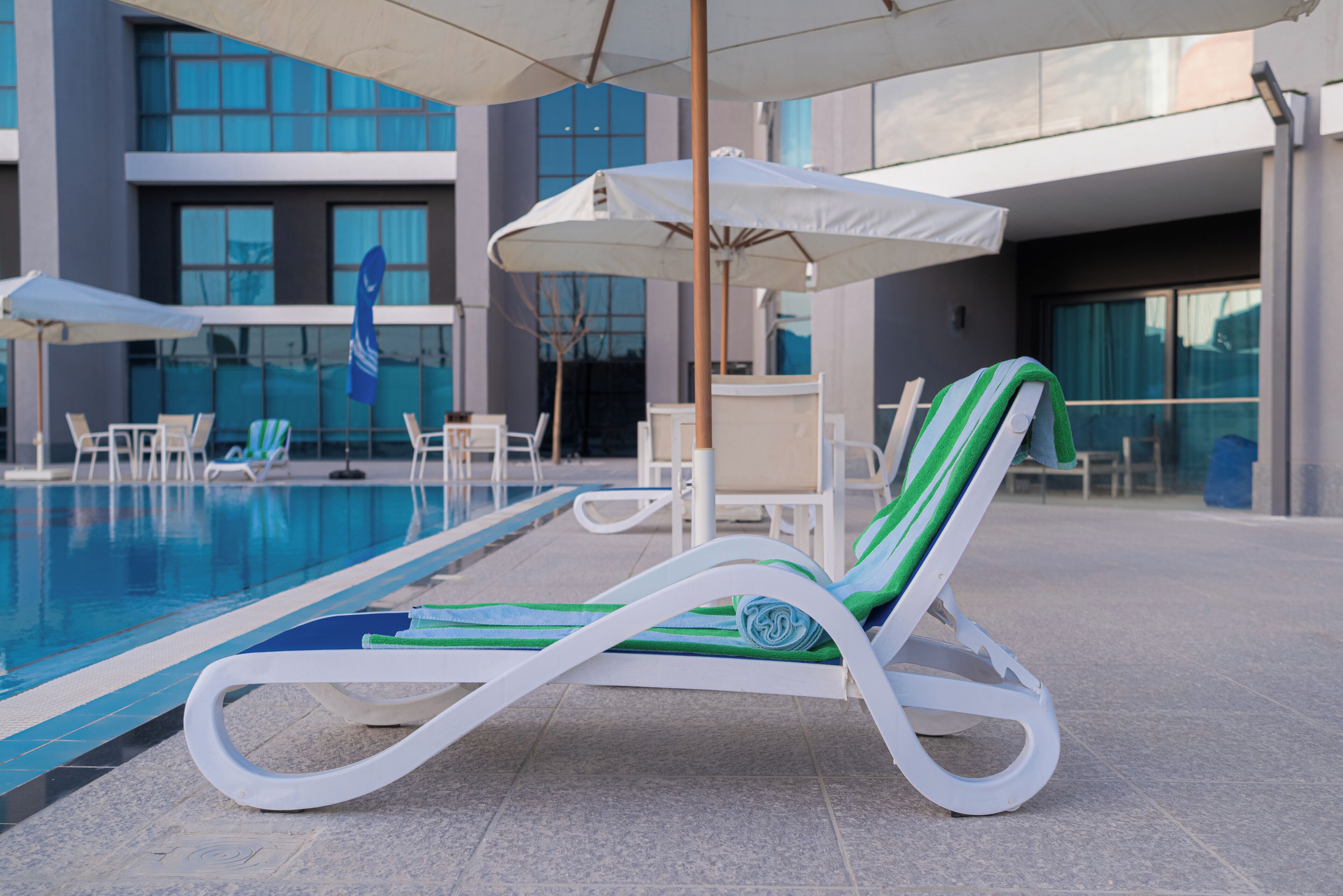 Zona junto a la piscina con sillones de descanso, sombrillas y un edificio moderno al fondo.