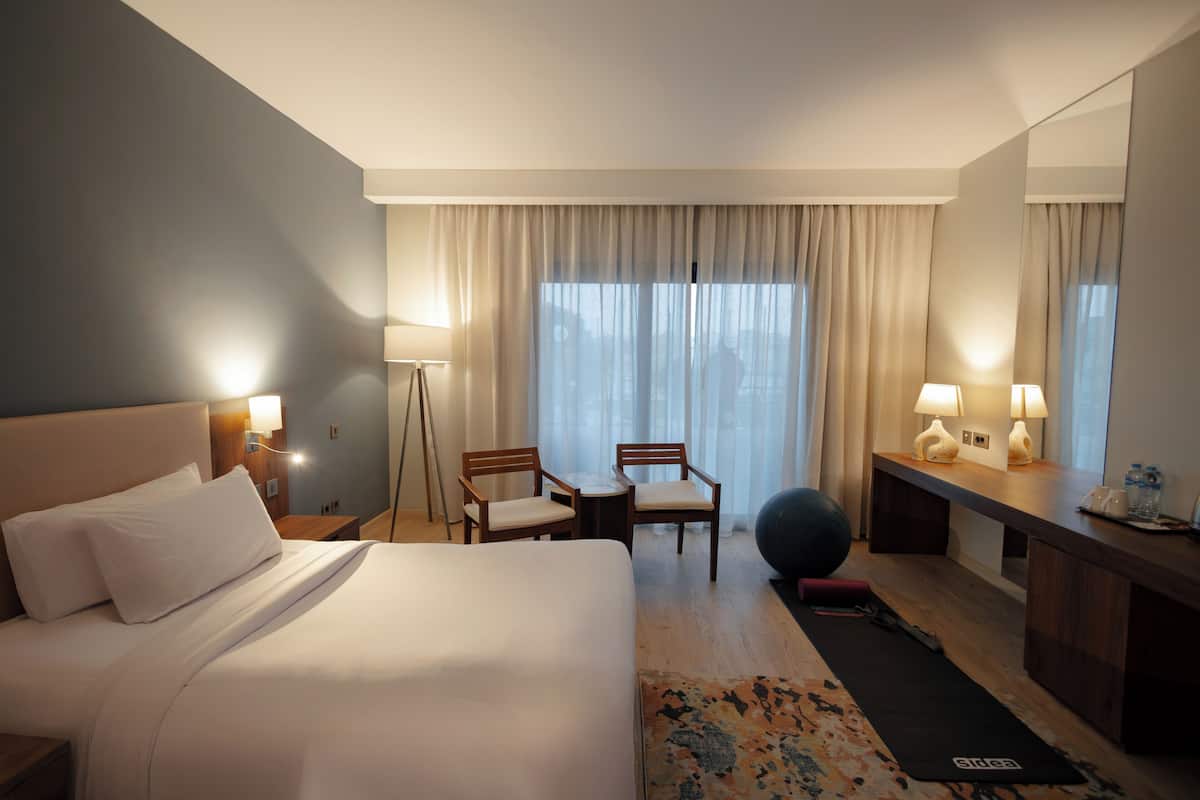 A hotel room with a bed, two chairs, a desk, a lamp, a large window with curtains, and a yoga mat.