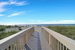 En träpromenad som leder till en strand med blå parasoller och en klar himmel.