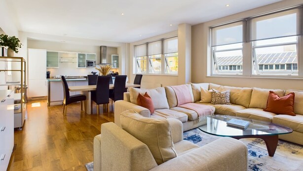 A modern living room with a beige sofa, a glass coffee table, and a dining area with a kitchen in the background. - Generous Covent Garden Apartment (London)