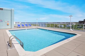 A rectangular swimming pool with clear blue water, surrounded by a tiled deck. There are blue lounge chairs and a white fence in the background.