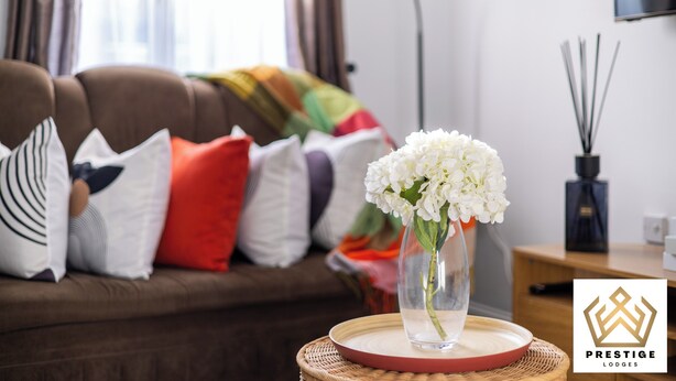 A living room with a brown sofa, colorful cushions, a vase with white flowers on a tray, and a Prestige Lodges logo.